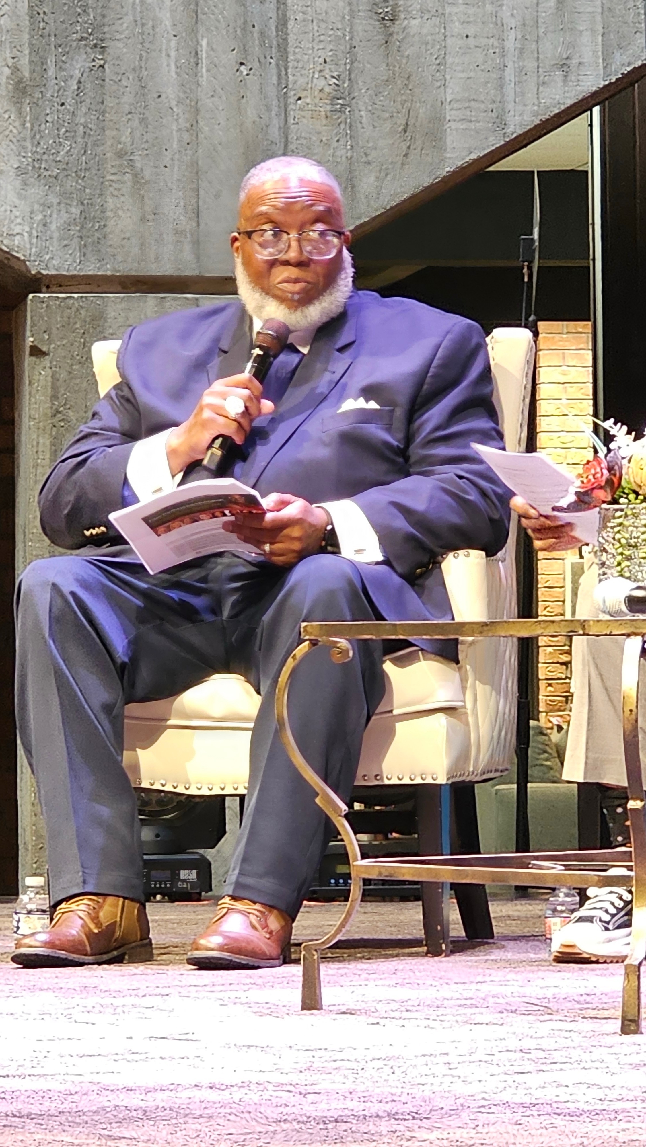 Dr. Preston T. Adams III, Senior Pastor and coach, seated mid-teaching in a navy suit holding a microphone.